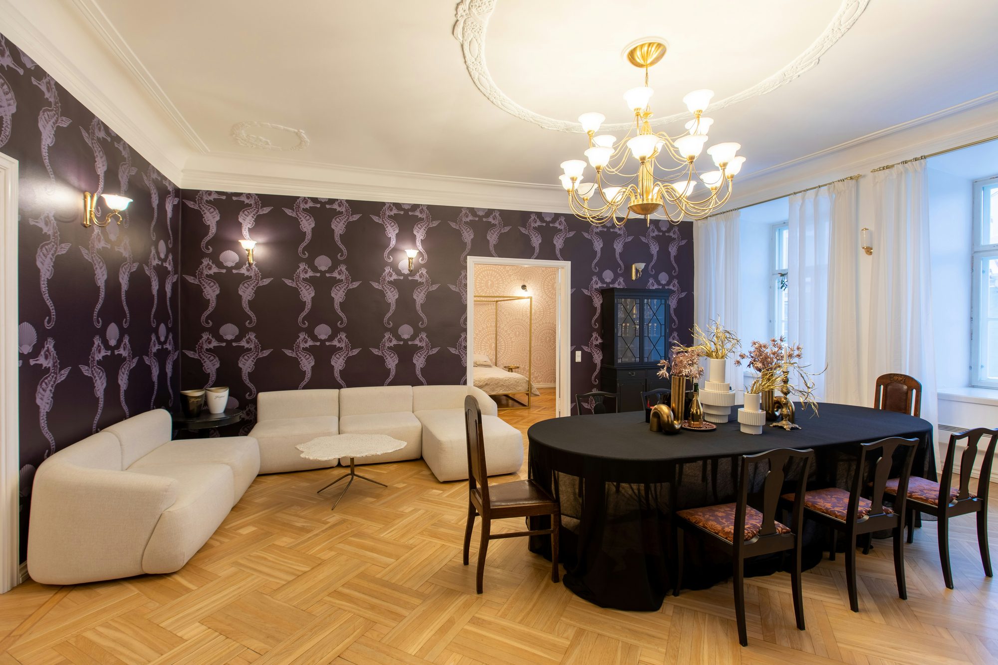Dining room with aubergine seahorse-pattern wallpaper, a brass chandelier, an ivory curved sofa and a long dark dining table on herringbone parquet