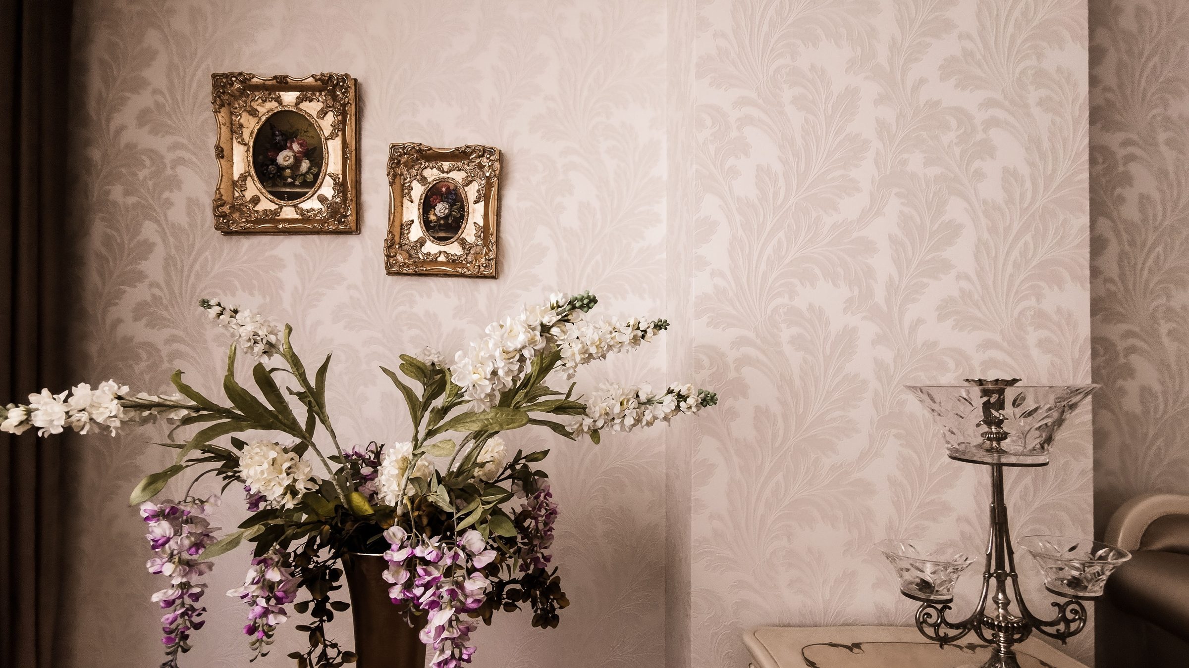 Close view of pale silver damask wallpaper with two gilt-framed miniatures, a vase of white and lilac flowers and a crystal epergne on a marble-topped table