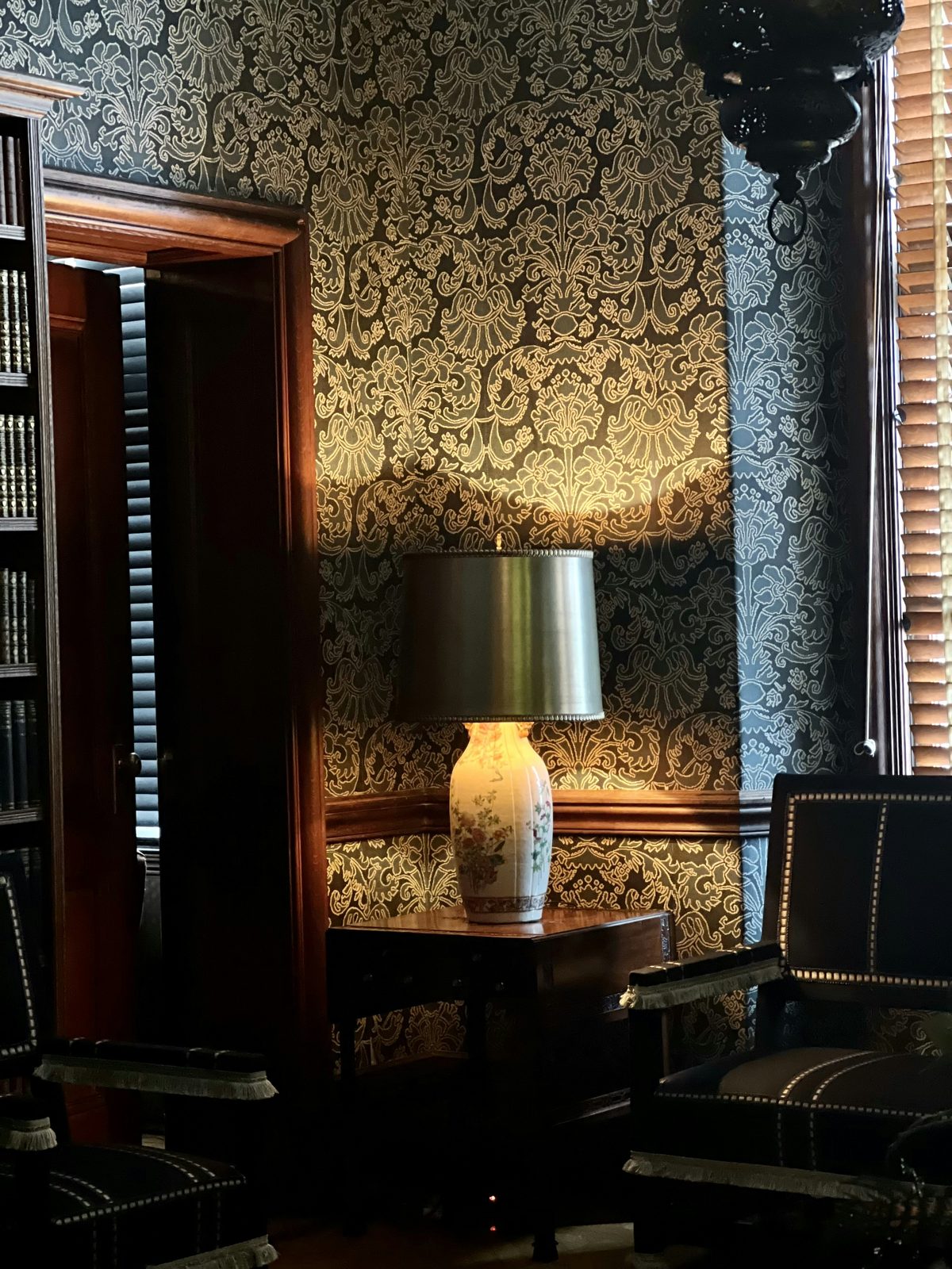Library corner with dark teal and gold damask wallpaper lit by a porcelain table lamp beside a dark wood doorway