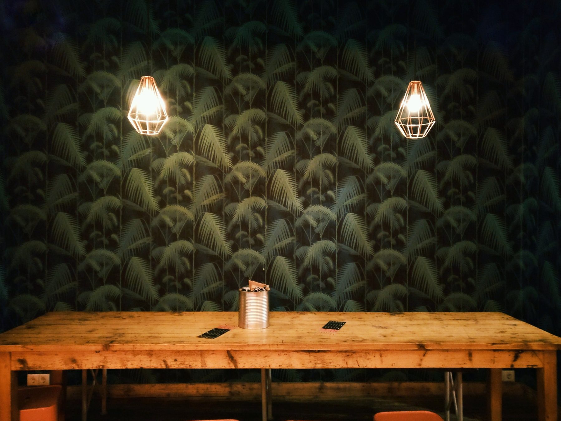 Dark palm-leaf wallpaper behind a long rustic wooden table under two copper cage pendant lights