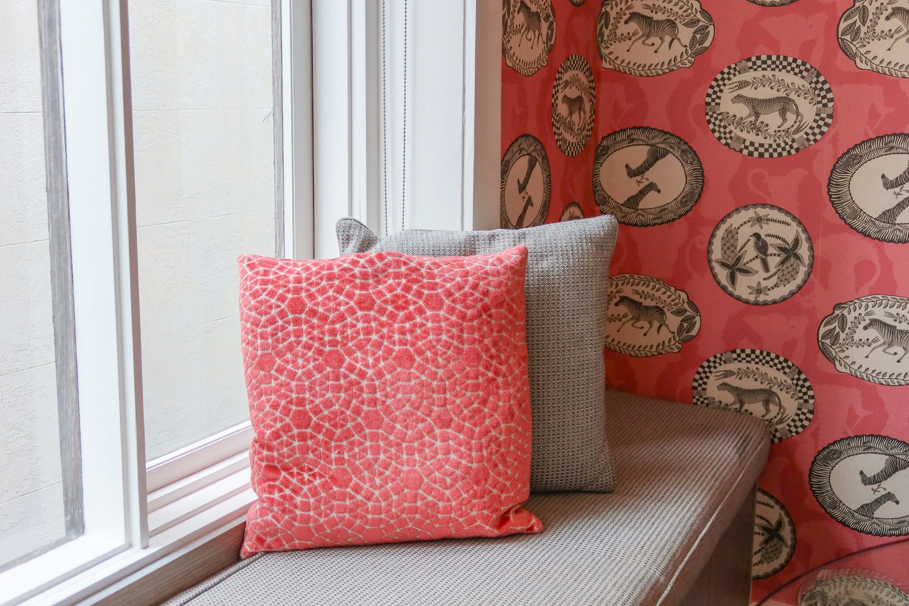 Window seat with coral and grey pillows against coral wallpaper printed with framed animal medallions