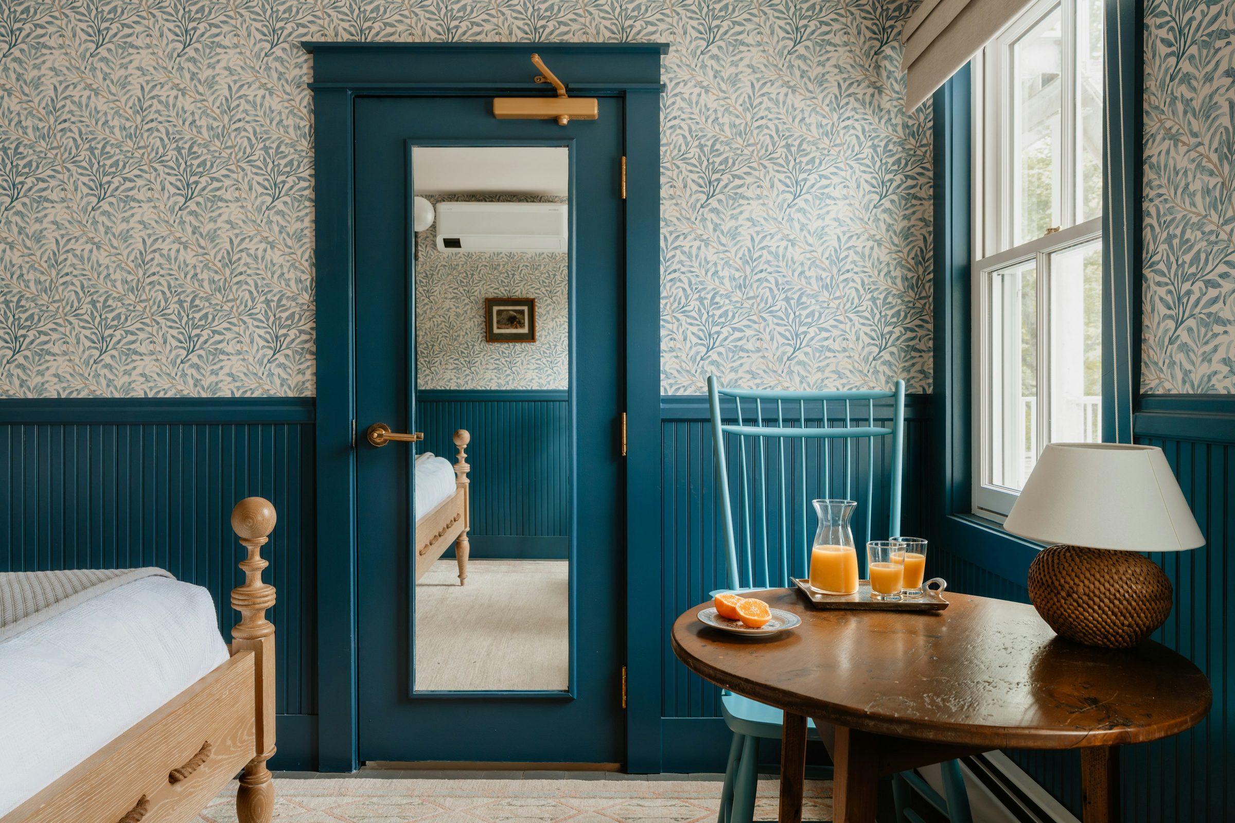 Bedroom corner with blue willow-leaf wallpaper above deep teal painted wainscoting, a mirrored teal door and a small wooden breakfast table by the window