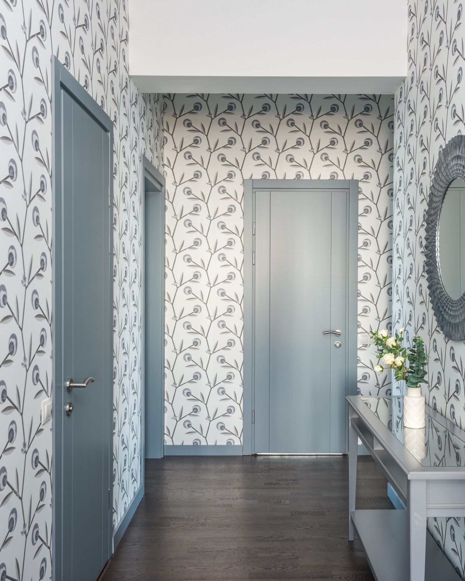 Bright hallway lined with white wallpaper printed with slate-blue botanical stems, with a round mirror, a grey console table and white flowers