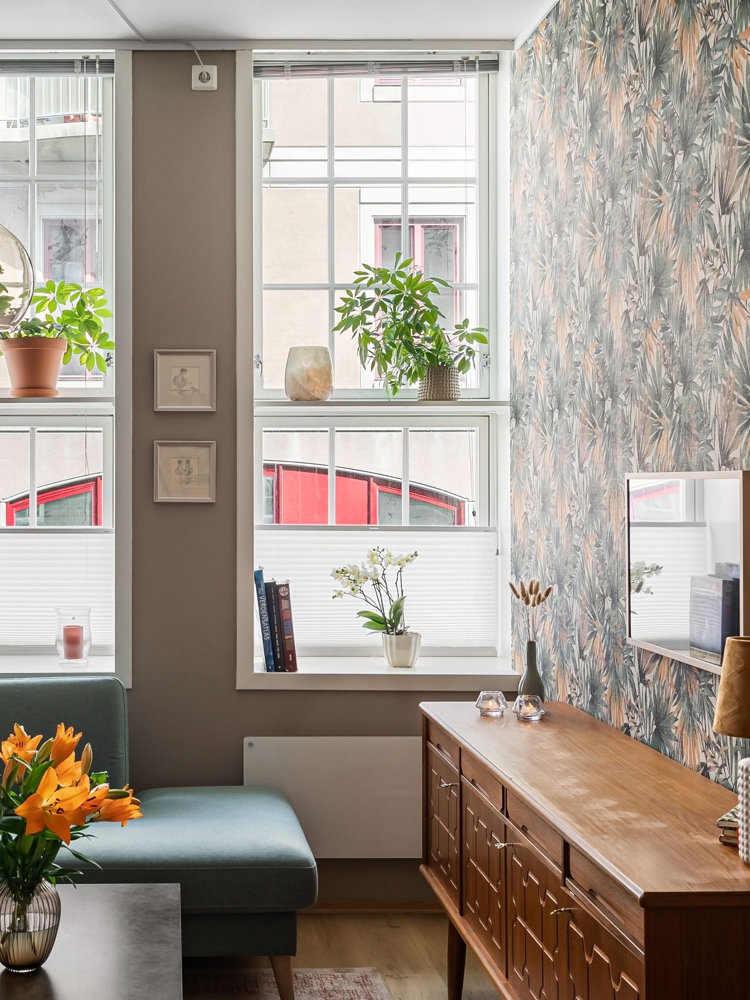Living room with a muted tropical-leaf wallpaper feature wall beside tall windows, a teal sectional sofa and a walnut sideboard